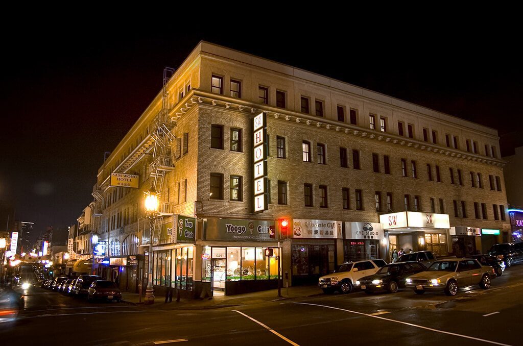 A nighttime view of a multi-story brick building on a street corner with cars parked along the street.