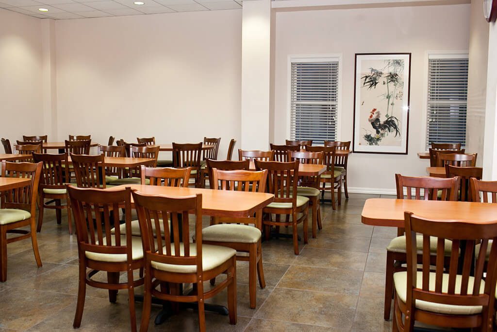A dining area with wooden tables and chairs, windows with blinds, and a large piece of artwork on the wall.
